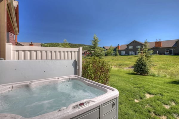 Hot tub on patio with view of green lawn, neighboring homes, and forested hillside under clear blue sky