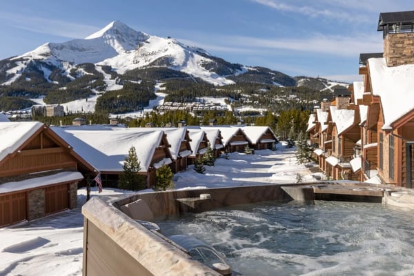 Snowy mountain resort with hot spring pool, wooden lodge buildings, and snow-covered peak in background
