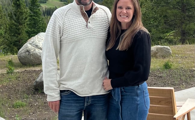 Couple standing on patio with snow-capped mountain and forested landscape behind them